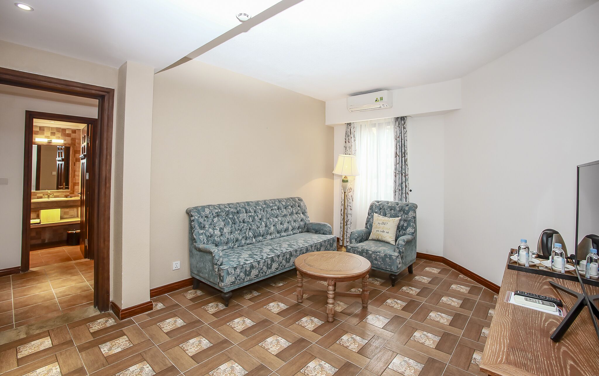 A hotel room with a patterned blue sofa, a matching armchair, a small wooden table, and a minibar.