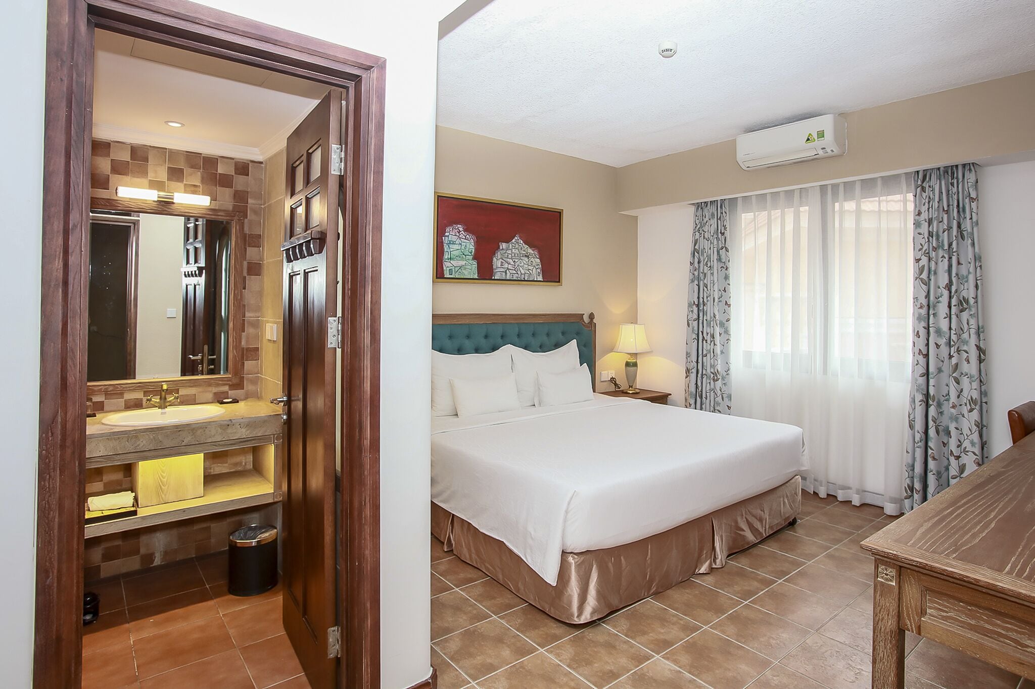 Habitación de hotel con una cama, una mesita de noche con lámpara, un baño con lavabo y espejo, y un cuadro en la pared.