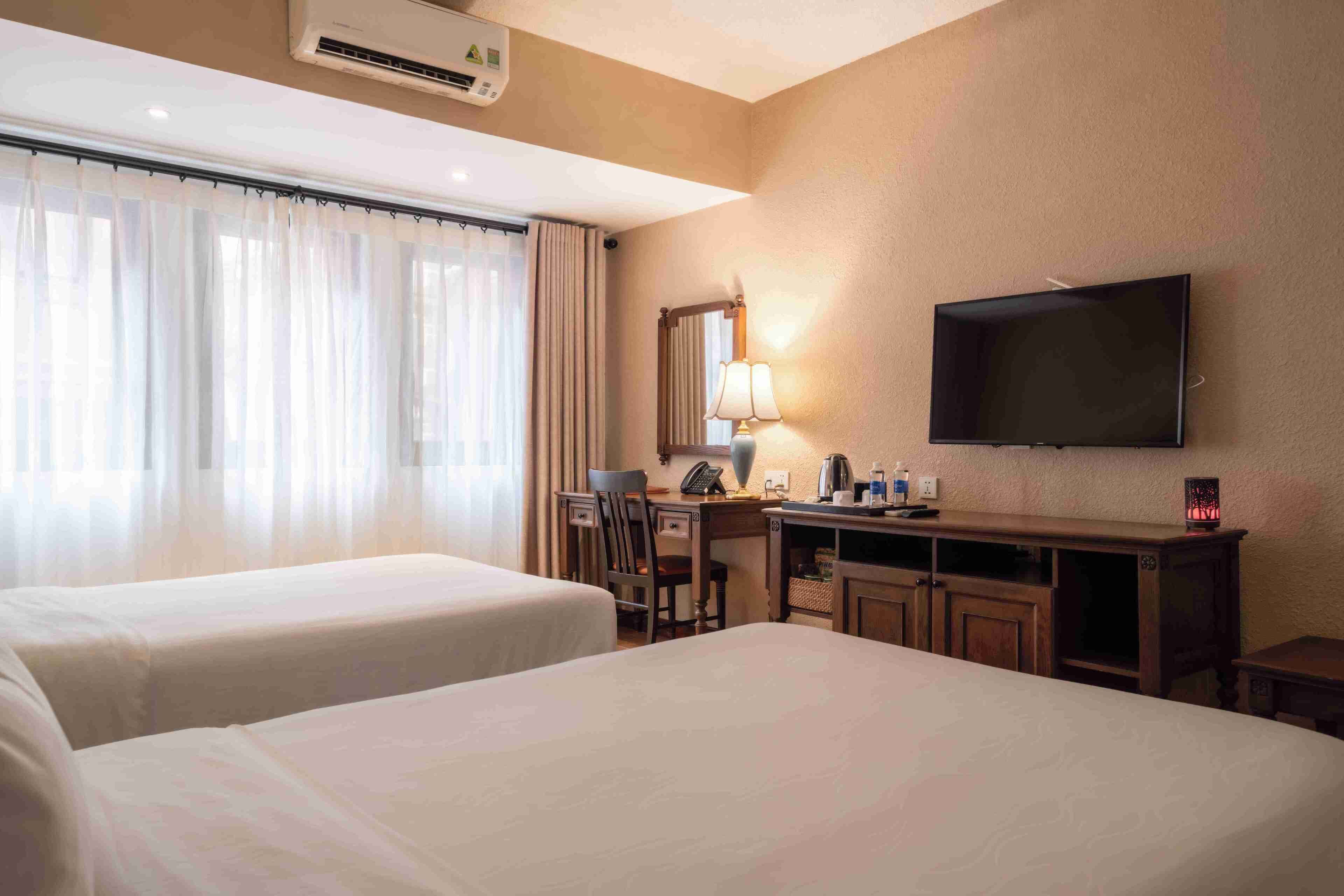 A hotel room with two beds, a desk with a lamp, a flat-screen TV, and a window with curtains.