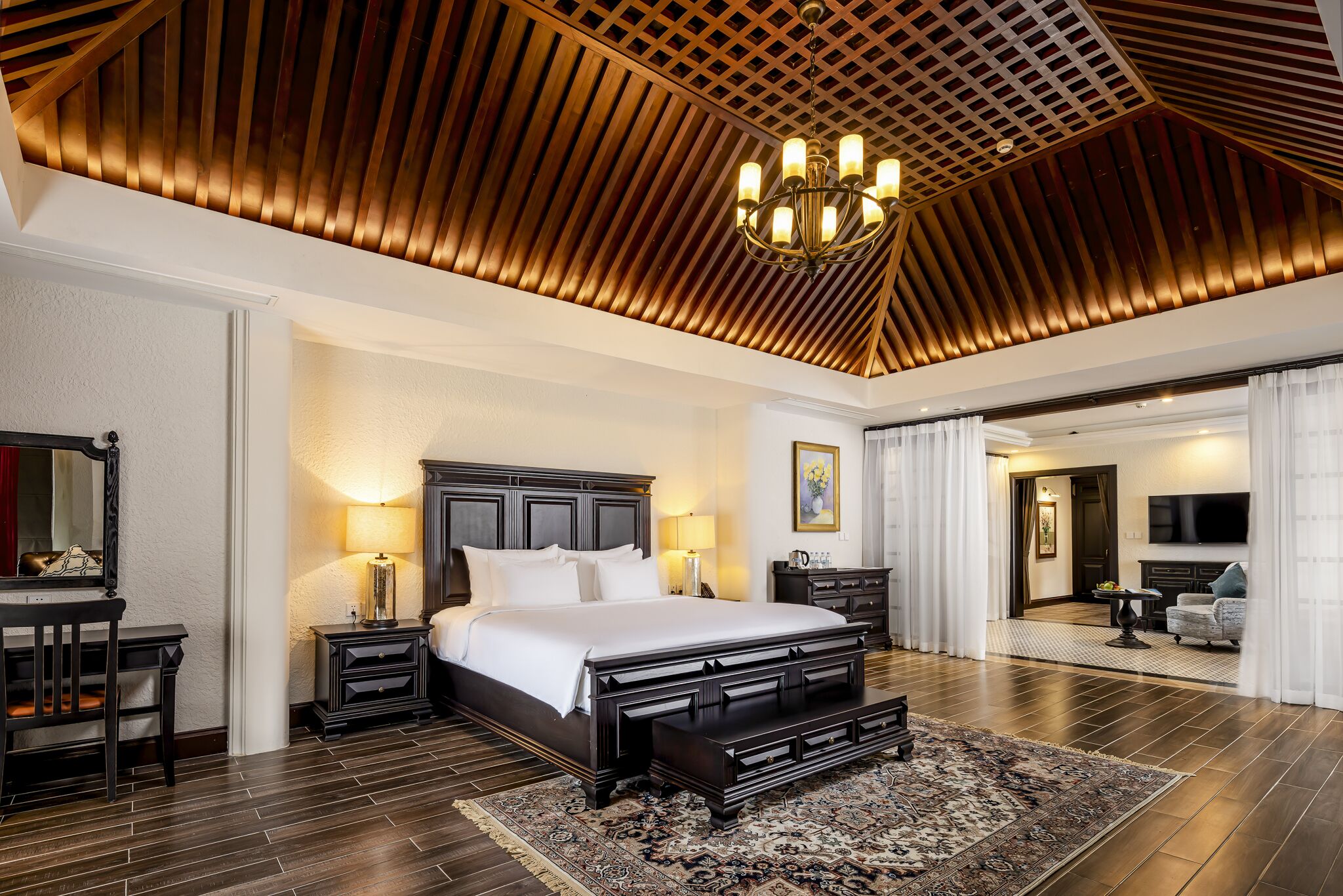 A spacious bedroom with a large bed, dark wood flooring, a chandelier, and a view into another room.