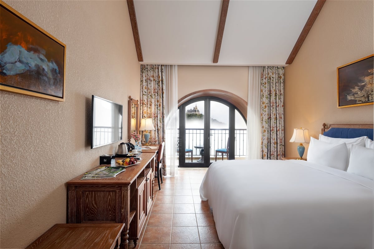 Une chambre d’hôtel avec un grand lit, un bureau avec un ordinateur, une télévision et une vue sur un château, visible à travers une fenêtre en arc de cercle.