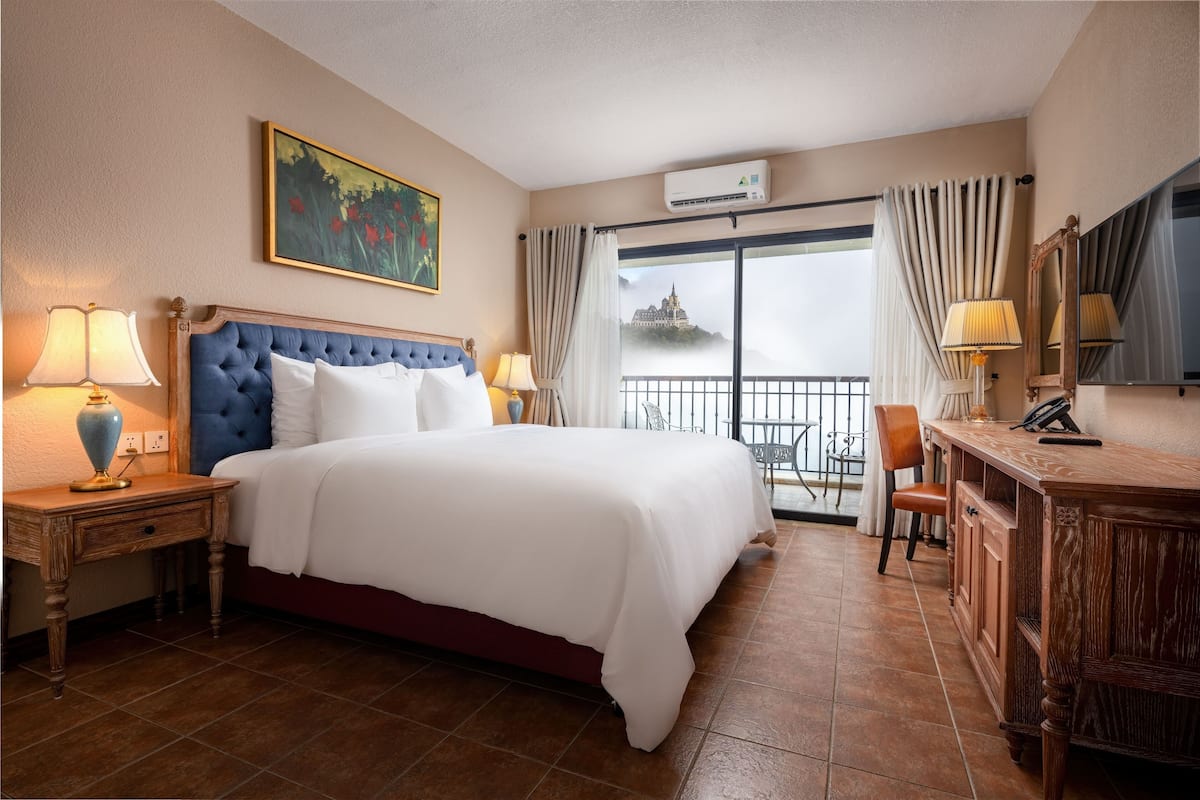 Une chambre d’hôtel avec un grand lit, un bureau, une chaise, un tableau et une vue sur un château par la fenêtre.