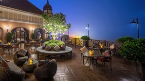 An outdoor patio with wicker furniture, tables, and chairs, surrounded by plants and a view of a building with a dome. - Venus Hotel Tam Dao (Tam Dao)