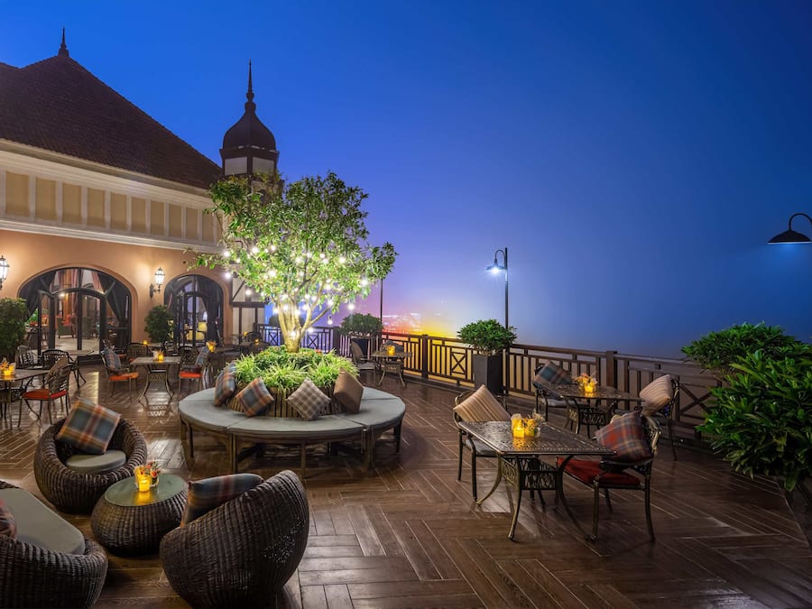Eine Terrasse im Freien mit Korbmöbeln, Tischen und Stühlen, umgeben von Pflanzen und Blick auf ein Gebäude mit einer Kuppel.