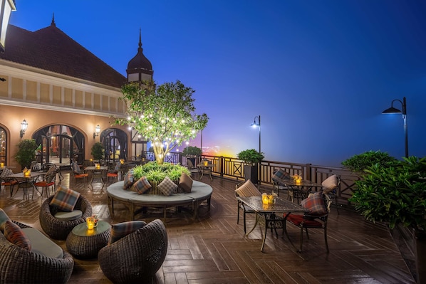 An outdoor patio with wicker furniture, tables, and chairs, surrounded by plants and a view of a building with a dome.