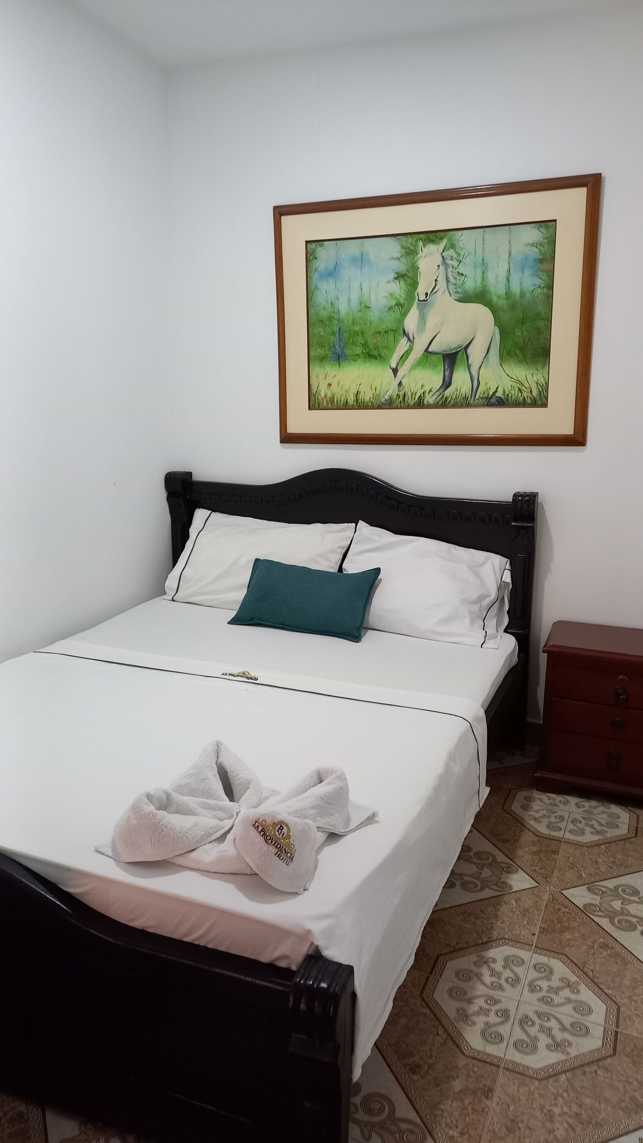 Una cama bien hecha con almohada blanca y aguamarina, un cuadro enmarcado de un caballo en la pared y una mesita de noche de madera al lado de la cama.