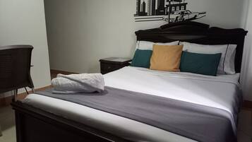 A neatly made bed with a white comforter, a gray blanket, and a folded white towel. There is a black headboard with a cityscape mural, a small bedside table with a lamp, and a chair with a woven backrest.