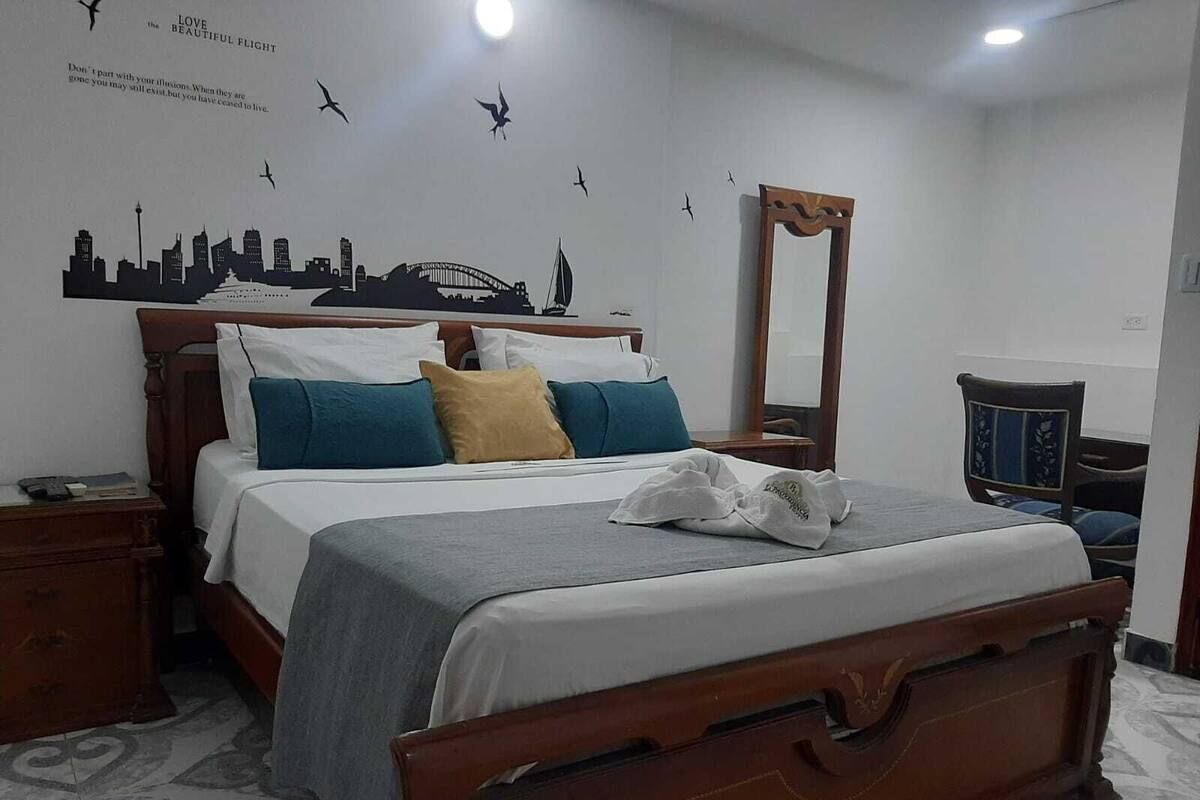 Un lit bien fait, avec une tête de lit en bois, une table de chevet avec une lampe et un mur orné d’une fresque représentant un paysage urbain.