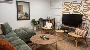 Una sala de estar con un sofá verde, una mesa redonda, sillas de mimbre, un televisor de pantalla plana y un panel decorativo en la pared.