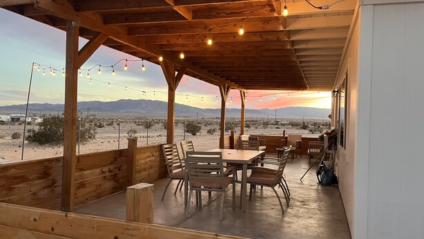 Un patio al aire libre con mobiliario de madera, luces colgantes y vistas a un paisaje desértico.