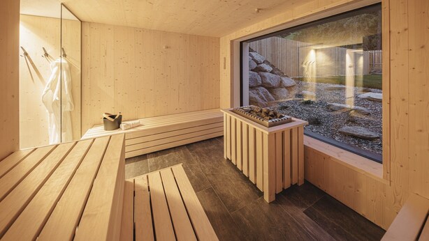 A wooden sauna room with a bench, a small table with stones, and a large window with a view of rocks and a wooden structure.