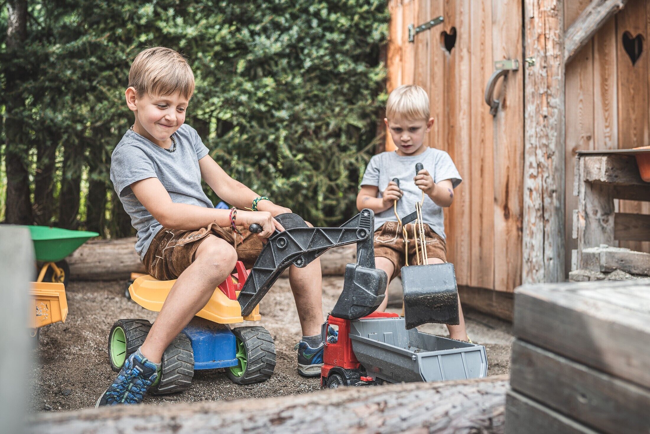 Two children playing with toy construction vehicles in an outdoor setting with a wooden shed in the background.