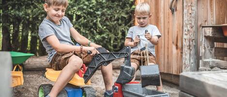 Two children playing with toy construction vehicles in an outdoor setting with a wooden shed in the background.