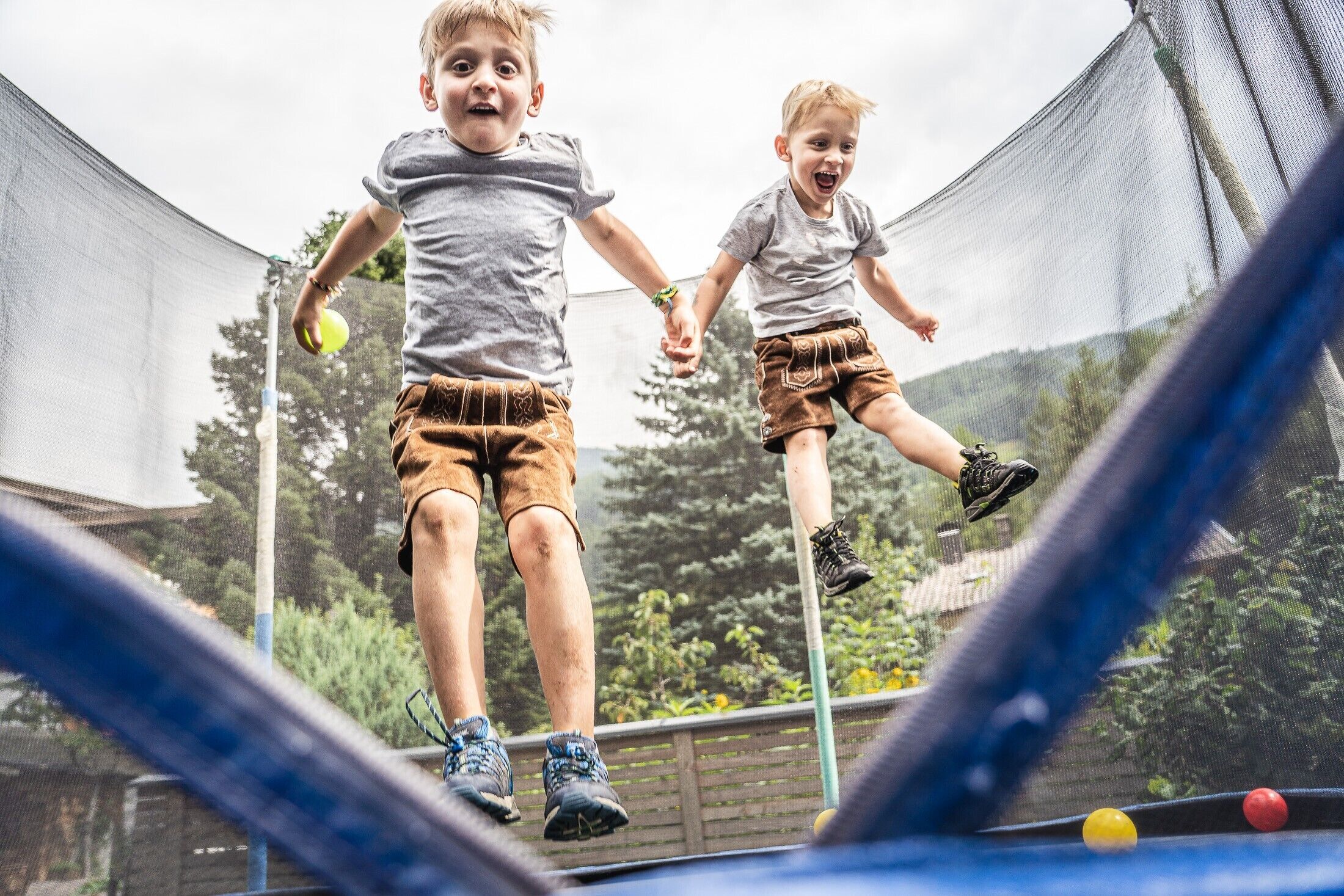 Two children playing on a trampoline with a netted enclosure, surrounded by greenery.