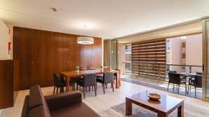 A modern living room with a sofa, dining table, and chairs, a wooden accent wall, and a view through a sliding door to an outdoor balcony.