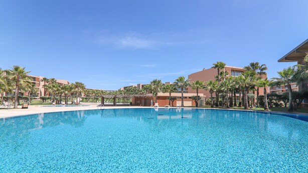 A large swimming pool with clear blue water, surrounded by palm trees and hotel buildings.