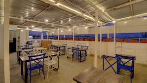 An indoor dining area with tables and chairs, a view of the cityscape through large windows, and a high ceiling with exposed beams and lighting fixtures.