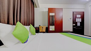 A hotel room with a bed, green pillows, a wooden nightstand, a mirror, a wardrobe, and a door with a notice.