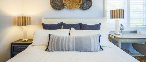 A neatly made bed with white bedding, a bedside table with a lamp, and a wall-mounted decorative basket.