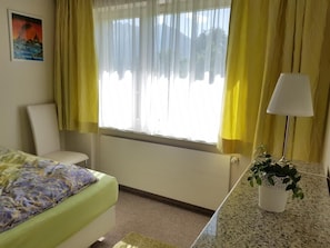 Une chambre d’hôtel avec un lit, une chaise, un bureau avec une lampe et une fenêtre avec des rideaux.
