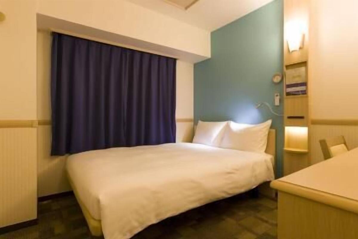 Hotellihuone, jossa on sänky, pieni työpöytä, ikkuna verhoilla ja seinälle kiinnitetty hylly, jossa on kello.