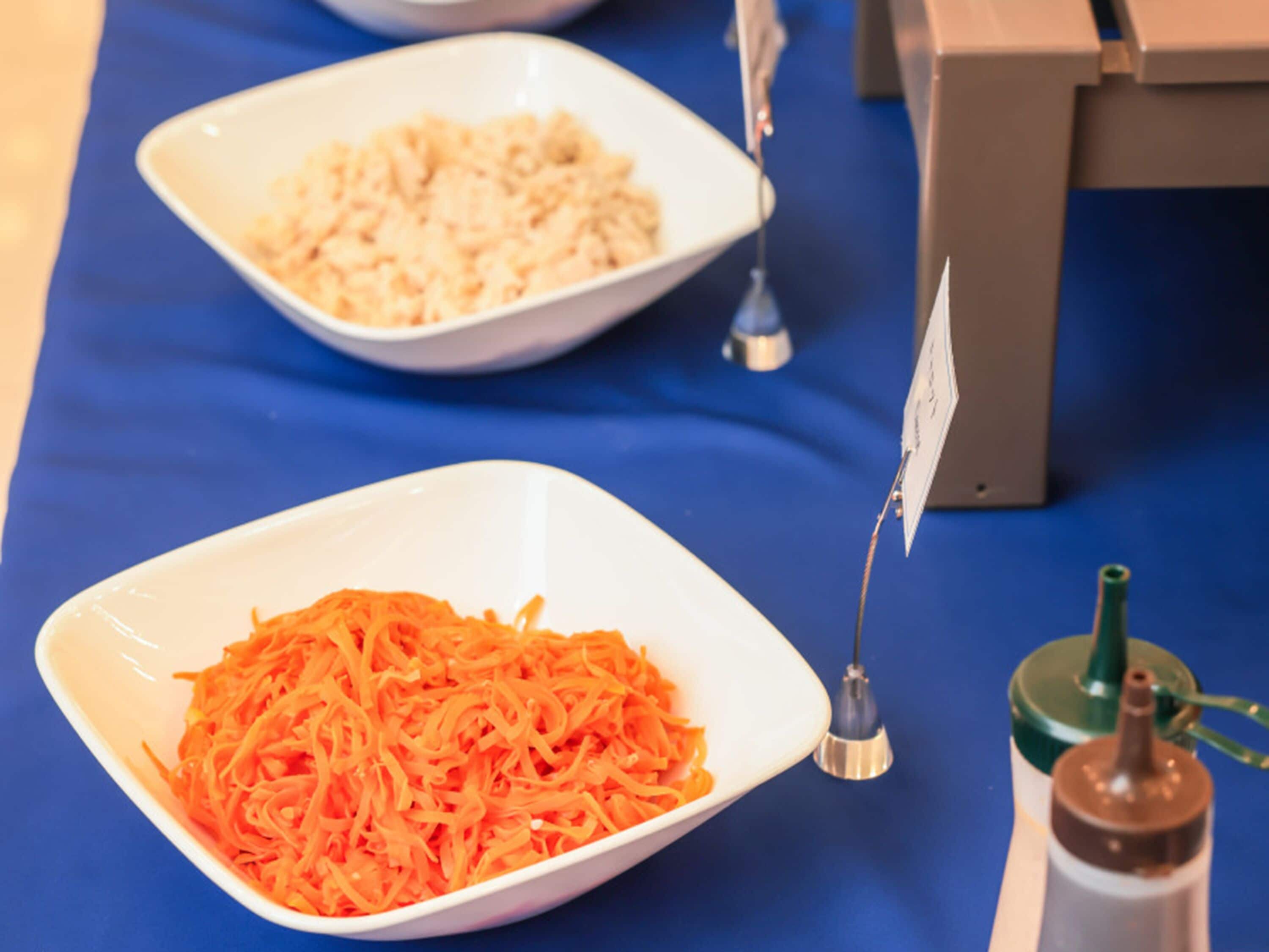Two white bowls on a blue tablecloth, one with shredded carrots and the other with a white substance.