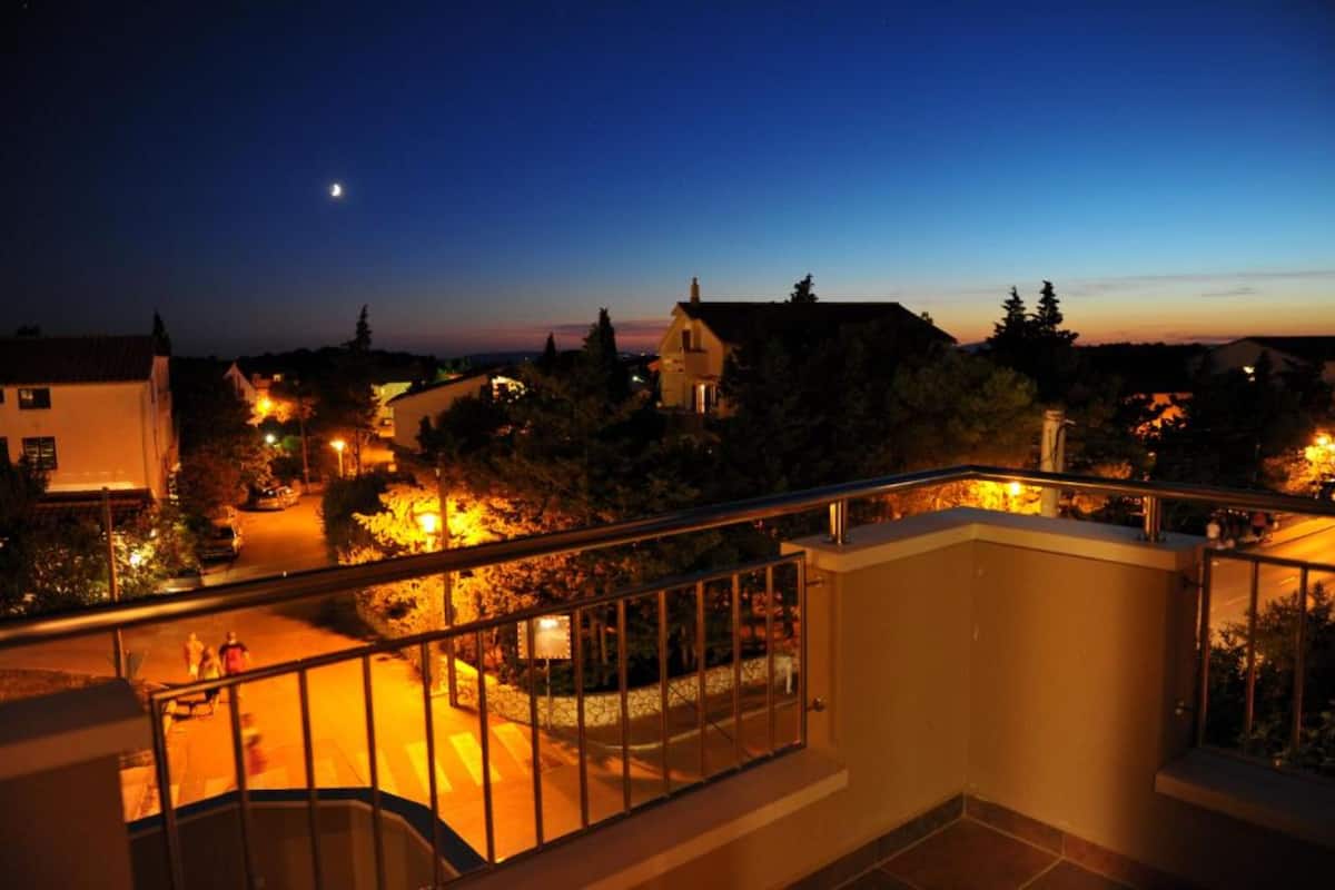 A nighttime view from a balcony overlooking a residential area with houses, a street, and a crescent moon in the sky.
