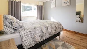 A bedroom with a bed, a nightstand, a lamp, a window with curtains, and a wall with framed artwork. - Lower Brambleside (Truro)