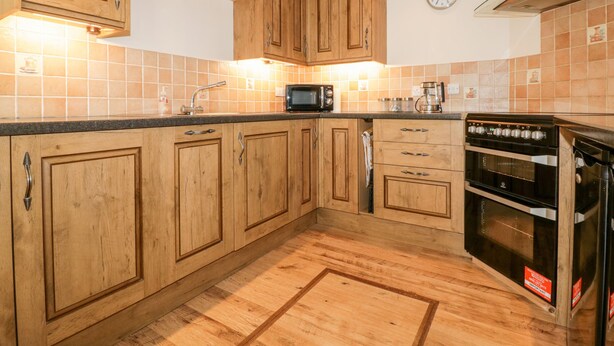 A kitchen with wooden cabinets, a microwave, a stove, and a clock on the wall. - Lower Brambleside (Truro)