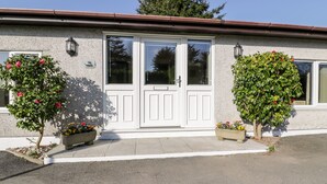 A building with a white door, two windows, and a small porch area. There are two potted plants with flowers on either side of the door. - Lower Brambleside (Truro)