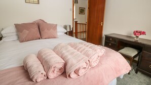 A neatly made bed with pink linens and rolled towels, a wooden desk with a vase of flowers, and a door leading to another room. - Thrushel Cottage (Lifton)