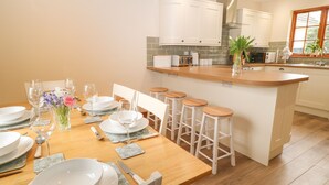 A modern kitchen with white cabinets, a wooden island with stools, and a dining area set for six. - Thrushel Cottage (Lifton)