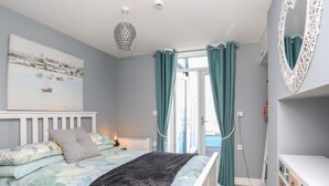 Um quarto com cama, mesa de cabeceira, luminária, janela com cortinas, espelho fixado na parede e um quadro.