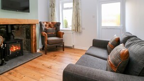 A living room with a fireplace, a sofa, a chair, and a television on the wall. - Gairnlea Cottage (Ballater)
