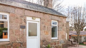 A stone building with a white door, windows, and a small porch area with outdoor furniture. - Gairnlea Cottage (Ballater)