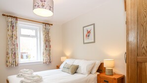 A neatly made bed with white linens, a wooden nightstand with a lamp, a window with floral curtains, and a framed picture on the wall. - Gairnlea Cottage (Ballater)