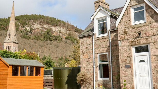 A stone house with a gabled roof, a small wooden structure to the left, and a church spire in the background. - Gairnlea Cottage (Ballater)