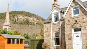 A stone building with a white door and windows, a wooden shed, and a church steeple in the background. - Gairnlea House (Ballater)