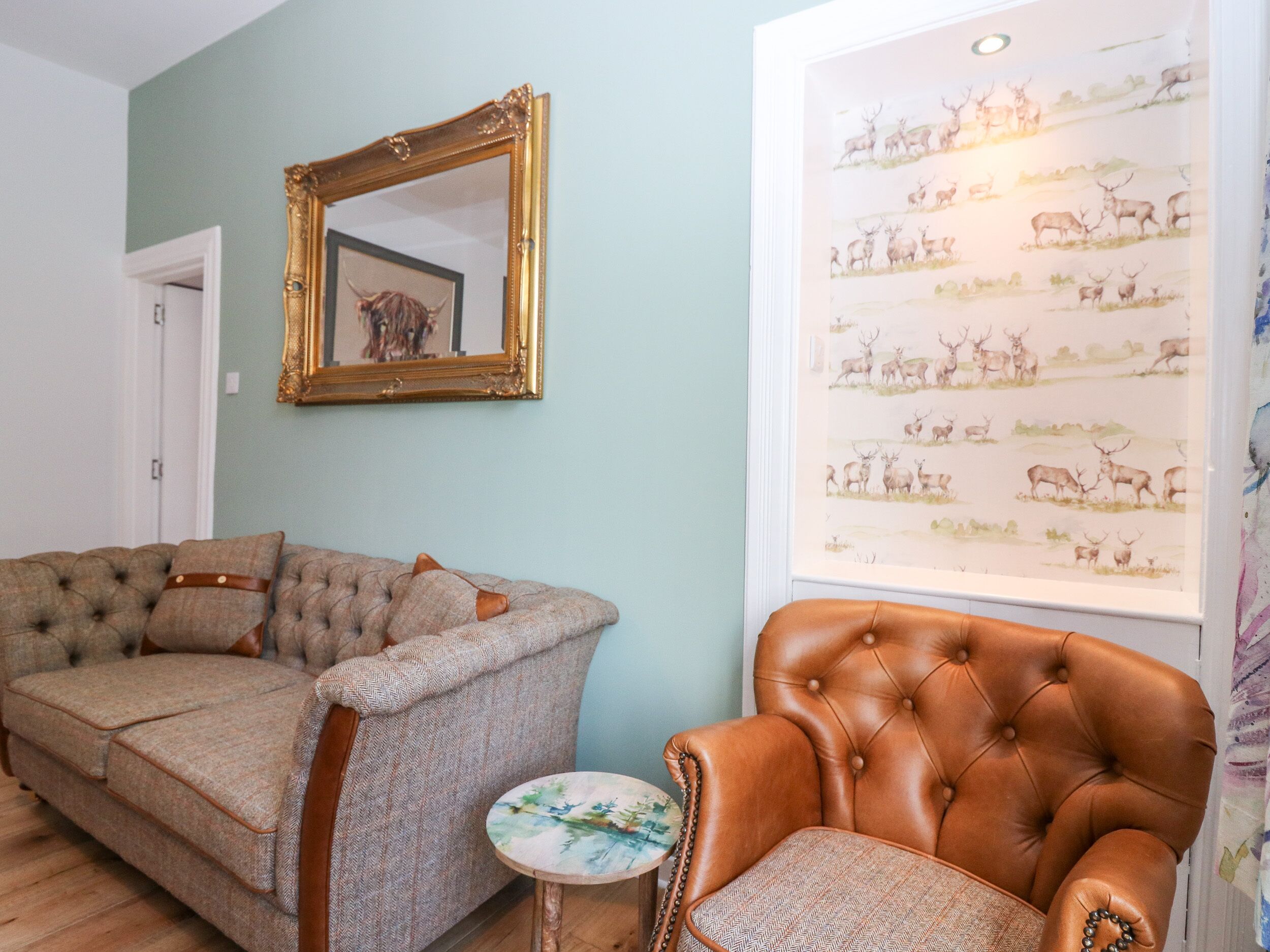 Un salon comprenant un canapé capitonné, un fauteuil en cuir, un miroir encadré et un mur orné d’une image représentant des animaux sauvages.