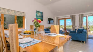 A dining area with a wooden table set for four, surrounded by wooden chairs. A vase with flowers is on the table, and there is a view of the outdoors through large windows. - Mellydonen Beder Del , Tor Down Quarry (Bodmin)