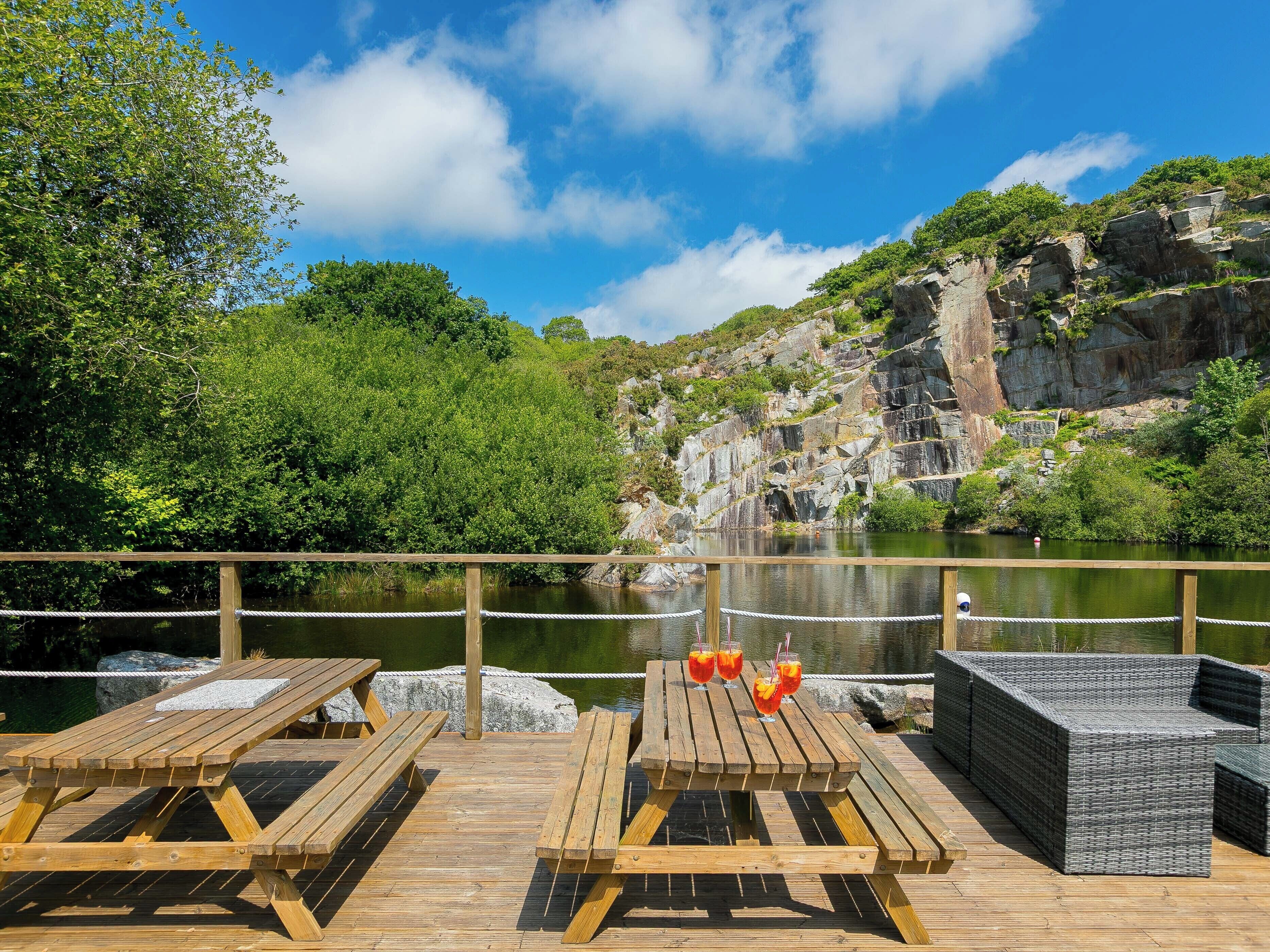 A wooden deck with picnic tables and wicker furniture overlooking a calm body of water, with a rocky cliff and greenery in the background.