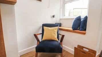Une chaise avec un coussin jaune, un banc en bois avec des coussins bleus et une fenêtre donnant sur des immeubles.