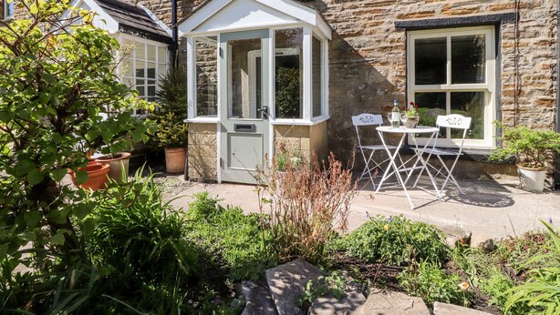 Une maison en pierre avec une véranda, un jardin agrémenté de plantes en pot et une table avec des chaises à l’extérieur.