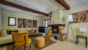 Een woonkamer met een bank, gele stoelen, een televisie, een open haard en kunstwerken aan de muur.