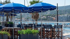 Buiten zitplaatsen met blauwe parasols, houten tafels en stoelen, met uitzicht op een jachthaven met boten en gebouwen op de achtergrond.