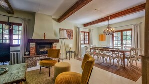 Een ruime woonkamer met een open haard, houten balken, een eethoek met witte stoelen en grote ramen.