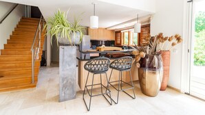 A modern kitchen with wooden cabinets, a central island, bar stools, and a large potted plant.