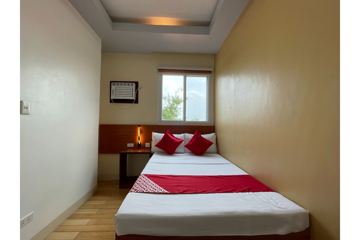 A small, narrow hotel room with a single bed, a nightstand, a window, and a ceiling fan.