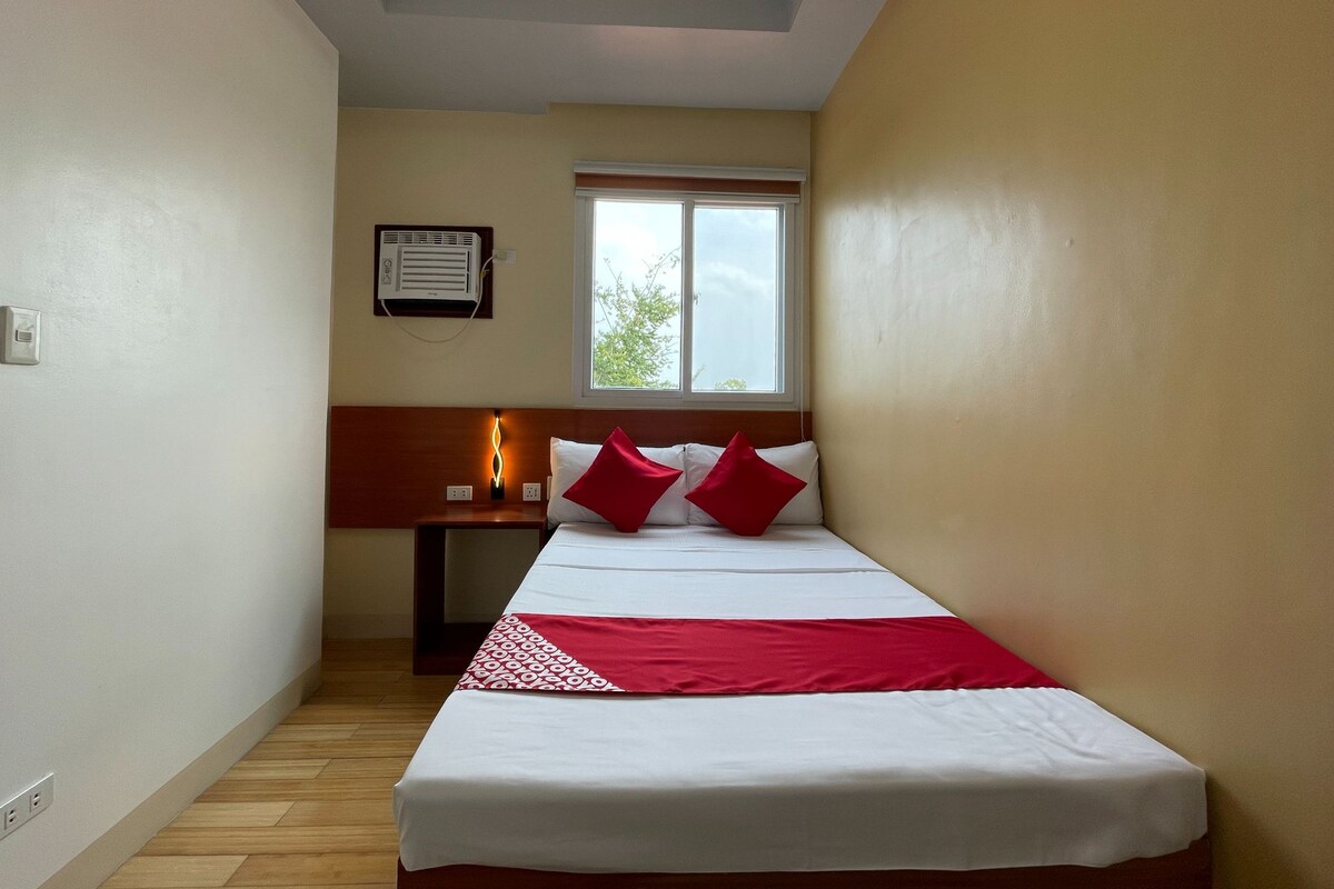 Una camera d'albergo piccola e stretta con un letto singolo, un comodino, una finestra e una ventola a soffitto.