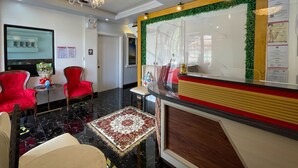 Um hall de hotel com balcão de recepção, cadeiras vermelhas e bege, um candelabro e um tapete estampado.
